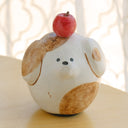 Clay sculpture of a round character, modeled to look like a dog without any arms or legs. Atop its head is an apple.