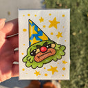 A hand holds a small artwork featuring a cartoon clown face with a yellow and blue party hat, green hair, a red nose, and yellow stars, photographed outdoors with pavement and grass in the background.