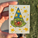 A hand holds a small hand-drawn cartoon clown face wearing a blue and yellow star-patterned hat, with yellow stars on a light background. The photo is taken outdoors with grass and pavement visible in the background