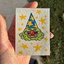 A hand holding a small artwork featuring a green cartoon face with a blue party hat and yellow stars, photographed outdoors with grass and pavement in the background.