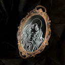 White engraved illustration on a mirror with gold ornate detailing on the exterior. Image is of a woman with long, flowing hair and a serious expression.