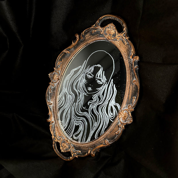 White engraved illustration on a mirror with gold ornate detailing on the exterior. Image is of a woman with long, flowing hair and a serious expression.