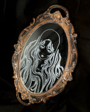 White engraved illustration on a mirror with gold ornate detailing on the exterior. Image is of a woman with long, flowing hair and a serious expression.