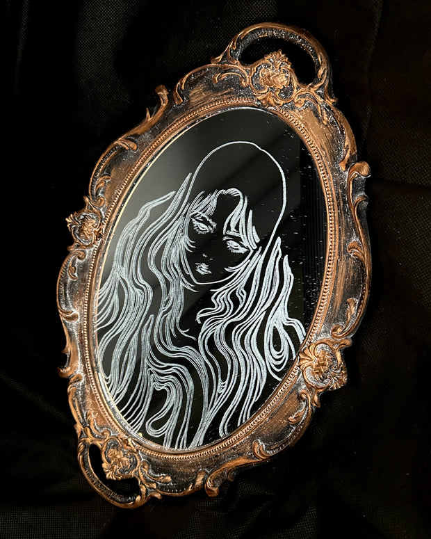 White engraved illustration on a mirror with gold ornate detailing on the exterior. Image is of a woman with long, flowing hair and a serious expression.