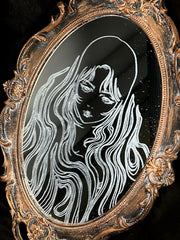 White engraved illustration on a mirror with gold ornate detailing on the exterior. Image is of a woman with long, flowing hair and a serious expression.