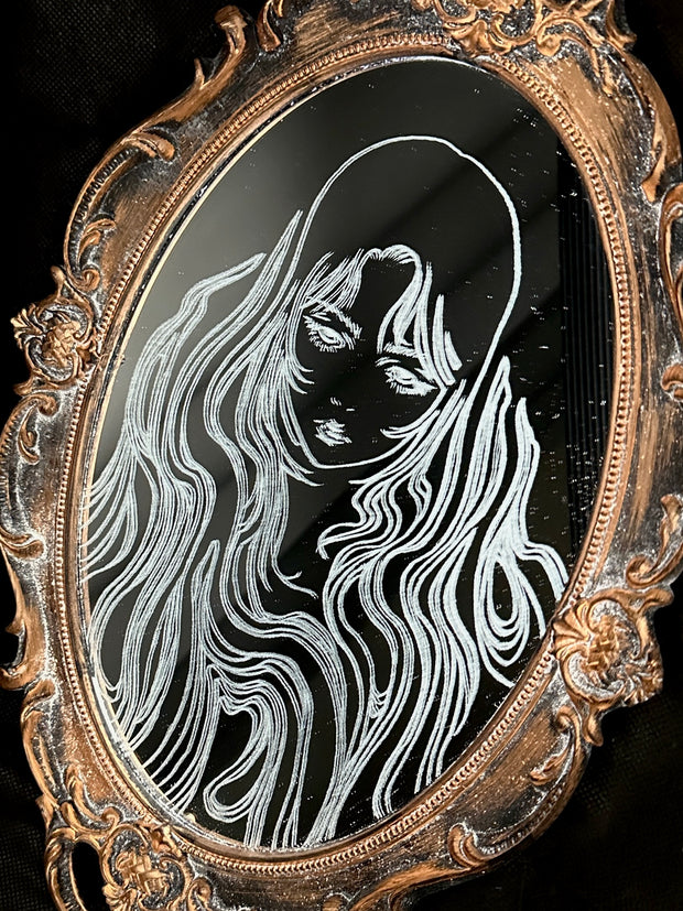 White engraved illustration on a mirror with gold ornate detailing on the exterior. Image is of a woman with long, flowing hair and a serious expression.