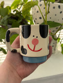 Cream colored mug with black spots and a cute, cartoon face to make it look like a stylized Dalmatian. Mug base is blue.