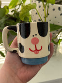 Cream colored mug with black spots and a cute, cartoon face to make it look like a stylized Dalmatian. Mug base is blue.