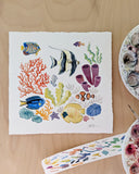 Watercolor illustration of several fish, sea plants and coral against a white background. Seen on a desk with paint palettes and test strips around it.