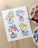 Watercolor illustration of colorful bonsai trees, all in colorful patterned pots and sprouting fruit. The piece is on a desk next to watercolor palettes. 