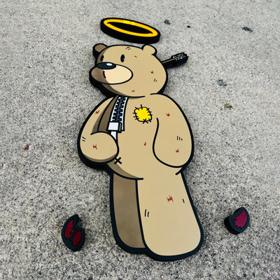 Die cut painted flat wood sculpture of a brown bear with a zipper down its front and a patch over its heart. It has an arrow in the back of its head and a halo overhead. Small red devil horns are on the ground nearby.