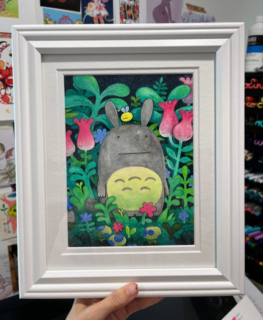 Illustration of a very stylized Totoro, with tiny dots for eyes, standing amongst colorful foliage. Framed and held in someone's hand.