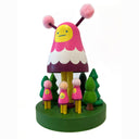 Colorful wooden toy figure with a pink hood and yellow face on a white background. It stands on a green platform with wooden trees and small figures that look like it.
