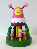 Colorful wooden toy figure with a pink hood and yellow face on a white background. It stands on a green platform with wooden trees and small figures that look like it.