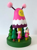 Colorful wooden toy figure with a pink hood and yellow face on a white background. It stands on a green platform with wooden trees and small figures that look like it.