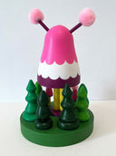Colorful wooden toy figure with a pink hood and yellow face on a white background. It stands on a green platform with wooden trees and small figures that look like it.