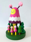 Colorful wooden toy figure with a pink hood and yellow face on a white background. It stands on a green platform with wooden trees and small figures that look like it.