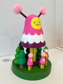 Colorful wooden toy figure with a pink hood and yellow face on a white background. It stands on a green platform with wooden trees and small figures that look like it.