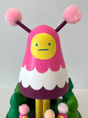 Colorful wooden toy figure with a pink hood and yellow face on a white background. It stands on a green platform with wooden trees and small figures that look like it.