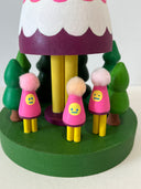 Colorful wooden toy figure with a pink hood and yellow face on a white background. It stands on a green platform with wooden trees and small figures that look like it.