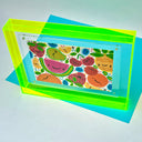 Painting of several colorful fruits with cartoon faces on them. Piece is framed in a fluorescent yellow color acrylic borderless frame.