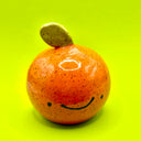 Small ceramic sculpture of an orange or a tangerine, round with a speckled finish and a cute cartoon smile drawn on. It has a single leaf coming out the top.