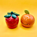 A pair of ceramic fruits - a tangerine, round with a speckled finish and a cute cartoon smile drawn on, and a strawberry with a similar cartoon happy face.