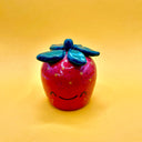 Ceramic sculpture of a strawberry with yellow dots and small speckling on its exterior, with a green leafy top and a cute, closed eye cartoon smile.