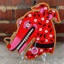 Piece of wood die cut in the shape of a dog's head with a long snout. The dog is painted bright red with pink and white flowers and has a cherry atop its nose.