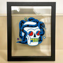 Painting atop of glass in an open back frame of a white skull with red eyes and yellow square teeth. Wrapped around the back and side of it is a blue snake with black patterning.