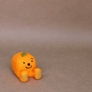 Small sculpture of a mandarin orange, with a simplistic smiling face and 2 feet, but no arms. It sits on the ground with its legs out in front and has a leaf atop its head.