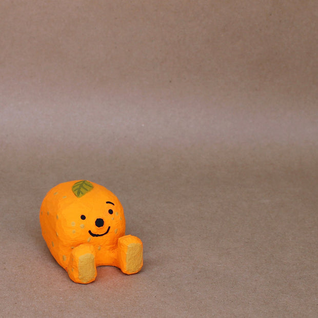 Small sculpture of a mandarin orange, with a simplistic smiling face and 2 feet, but no arms. It sits on the ground with its legs out in front and has a leaf atop its head.