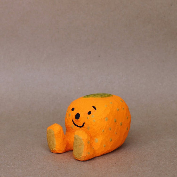 Small sculpture of a mandarin orange, with a simplistic smiling face and 2 feet, but no arms. It sits on the ground with its legs out in front and has a leaf atop its head.