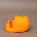 Small sculpture of a mandarin orange, with a simplistic smiling face and 2 feet, but no arms. It sits on the ground with its legs out in front and has a leaf atop its head. Viewed from the side.