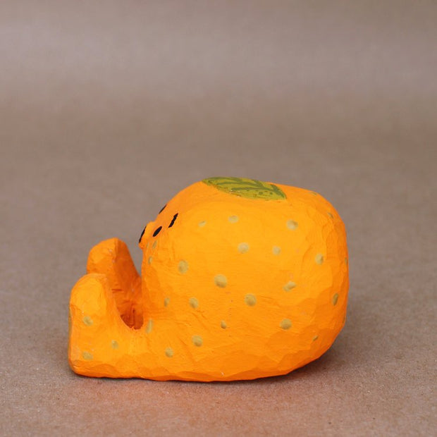 Small sculpture of a mandarin orange, with a simplistic smiling face and 2 feet, but no arms. It sits on the ground with its legs out in front and has a leaf atop its head. Viewed from the side.