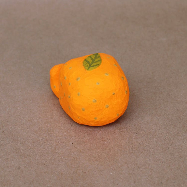 Small sculpture of a mandarin orange, with a simplistic smiling face and 2 feet, but no arms. It sits on the ground with its legs out in front and has a leaf atop its head. Viewed from the top.