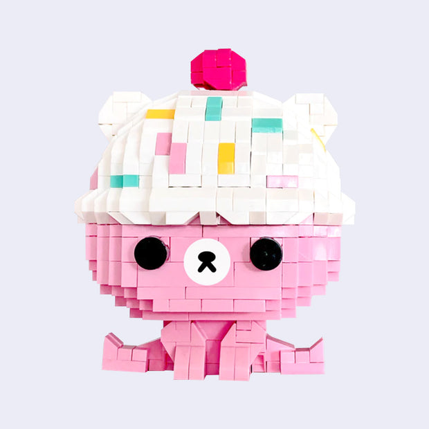 3D sculpture made out of small brick like plastic pieces of a pink bear sitting with its legs spread out. Its head is a white ice cream scoop with sprinkles and a cherry on top.