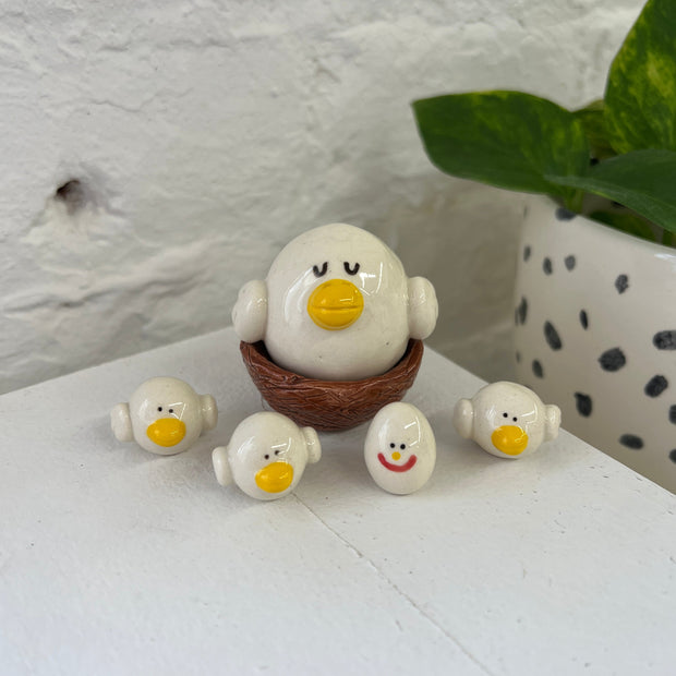 Ceramic sculpture of a white bird, resembling an egg with small wings and a yellow beak. It sits in a nest and around it are smaller birds and a single egg with a smiling face.