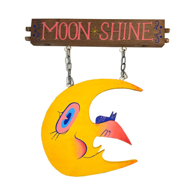 Die cut painted wooden sculpture of a crescent moon with a large curved nose and big red and blue eyes. It has a small blue cat on its nose and hangs from a post that reads "Moon Shine" in stylistic letters.