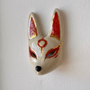 A hand-painted ceramic fox mask wall mask in beige with red and gold accents, mounted on a textured white wall.