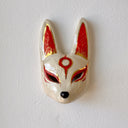 A hand-painted ceramic fox mask wall mask in beige with red and gold accents, mounted on a textured white wall.