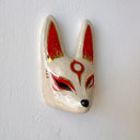 A hand-painted ceramic fox mask wall mask in beige with red and gold accents, mounted on a textured white wall.