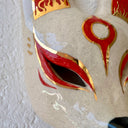 A hand-painted ceramic fox mask wall mask in beige with red and gold accents, mounted on a textured white wall.