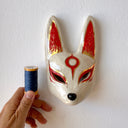 A hand-painted ceramic fox mask wall mask in beige with red and gold accents, mounted on a textured white wall.