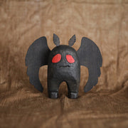 Sculpture of Mothman, the folklore legend, all black with a stout body and large flat wings. Its eyes are red and it has a simple disgruntled expression on its face.