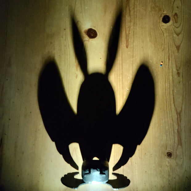 Sculpture of Mothman, the folklore legend, stylistically lit up from the back, casting a very large looming shadow despite its small size.