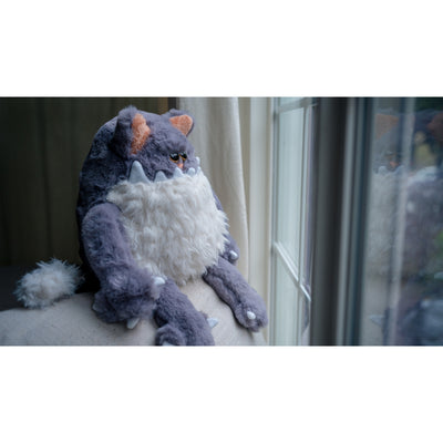 Fluffy plush sculpture of a gray cat, with a large jagged tooth underbite and a white fluffy belly that looks like a beard. It sits and looks off into the distance.