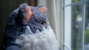 Fluffy plush sculpture of a gray cat, with a large jagged tooth underbite and a white fluffy belly that looks like a beard. It sits and looks off into the distance.