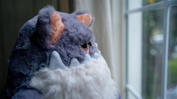 Fluffy plush sculpture of a gray cat, with a large jagged tooth underbite and a white fluffy belly that looks like a beard. It sits and looks off into the distance.