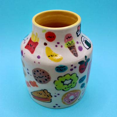 Ceramic vase with many small doodles on it of snacks, such as: fruit, fries, ice cream donuts and pie.